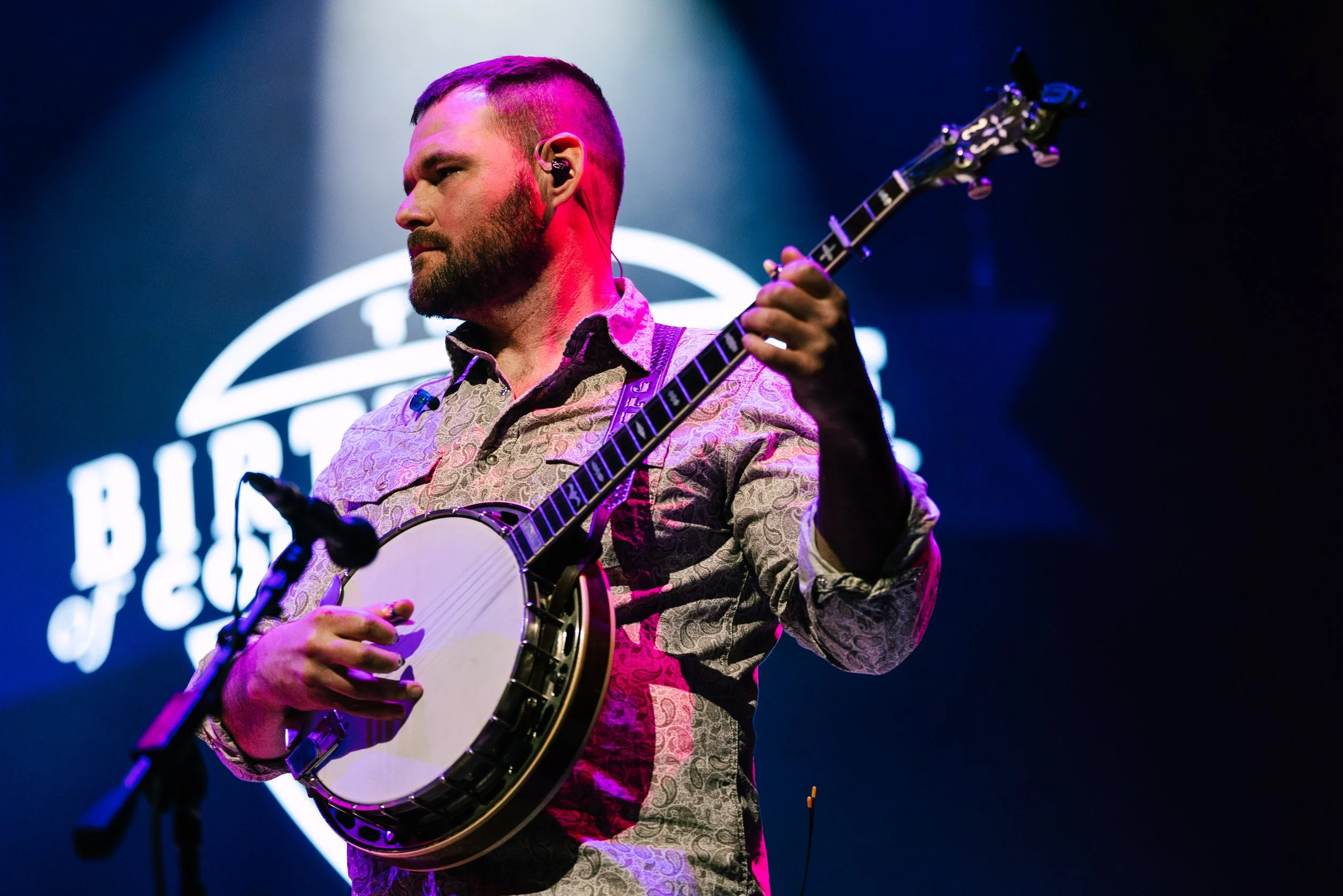A man with a beard and short hair playing a banjo on stage, illuminated by stage lights with a blurred logo in the background.