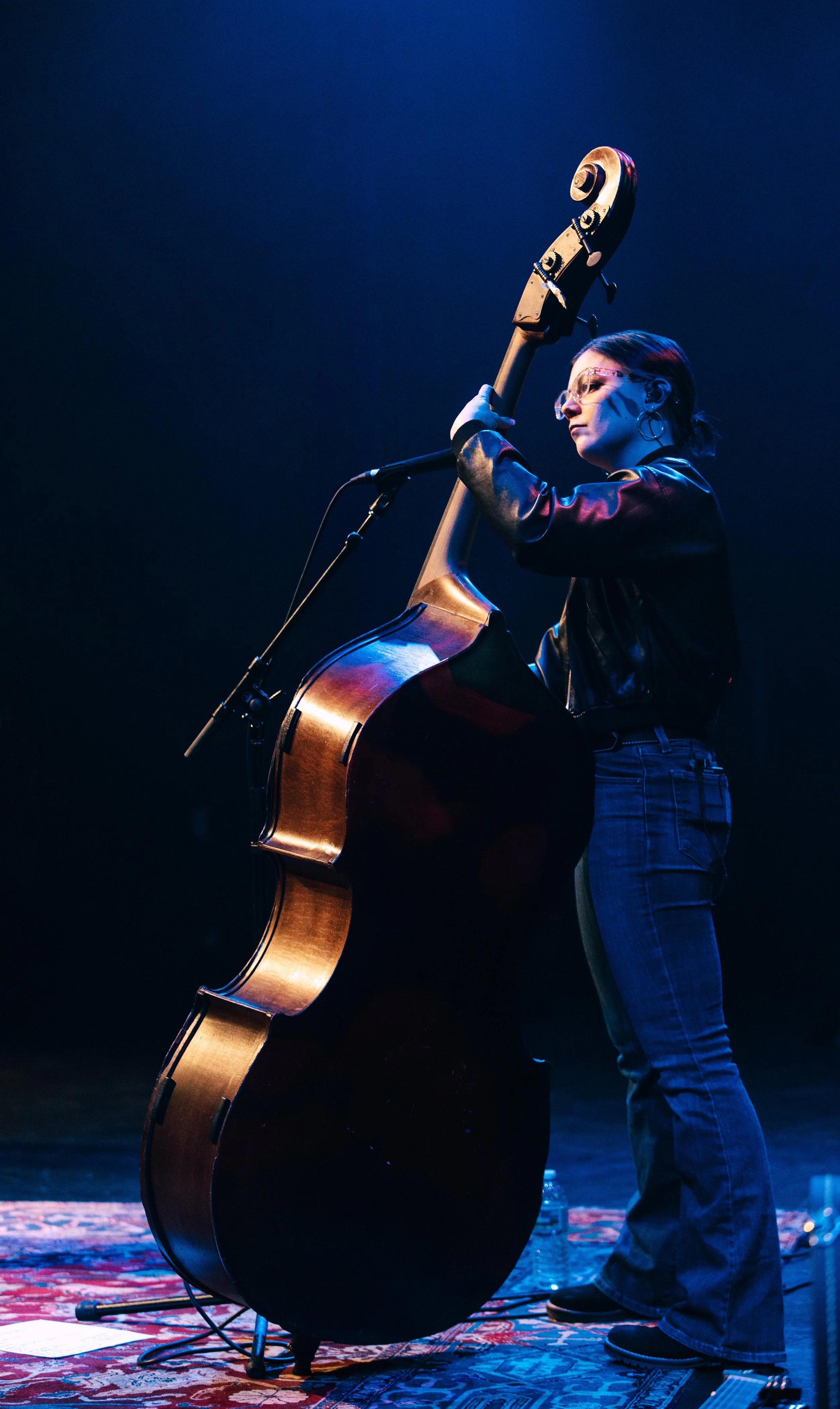 A woman playing a double bass on a stage with a microphone stand in front of her. She is wearing glasses, a black leather jacket, and jeans, and is looking to the side.