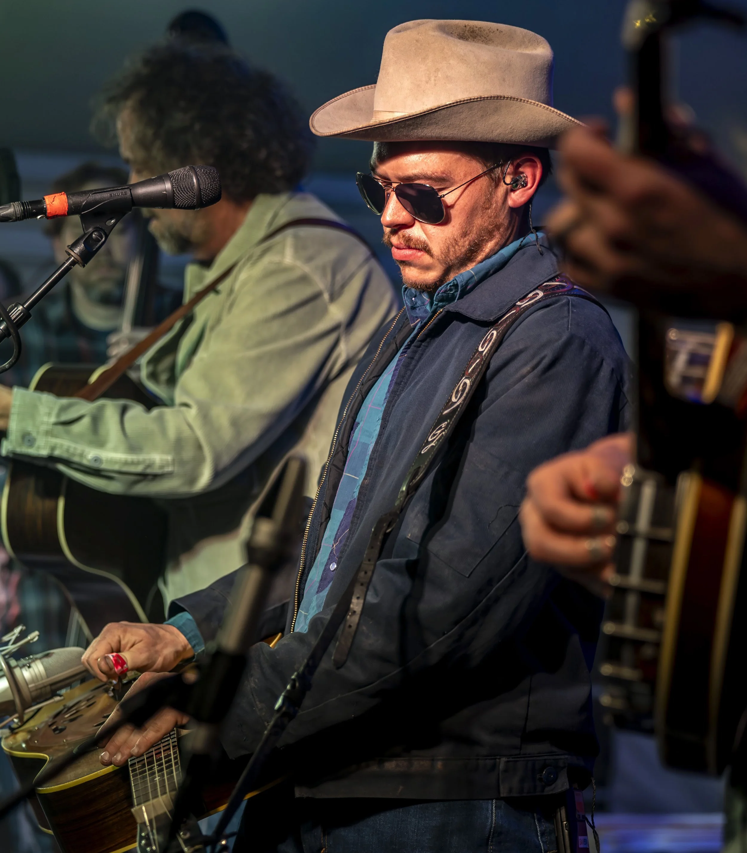 Musician wearing a wide-brimmed hat, sunglasses, and a blue jacket, playing an acoustic guitar at a performance.