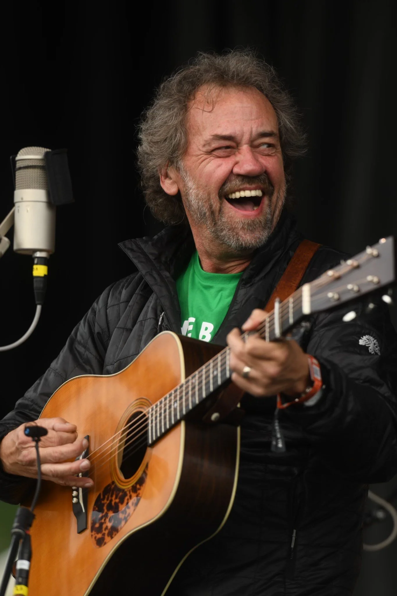 A man with curly, gray hair and a beard playing an acoustic guitar, singing or speaking into a microphone, wearing a black jacket and a green shirt, smiling and looking joyful against a black background.