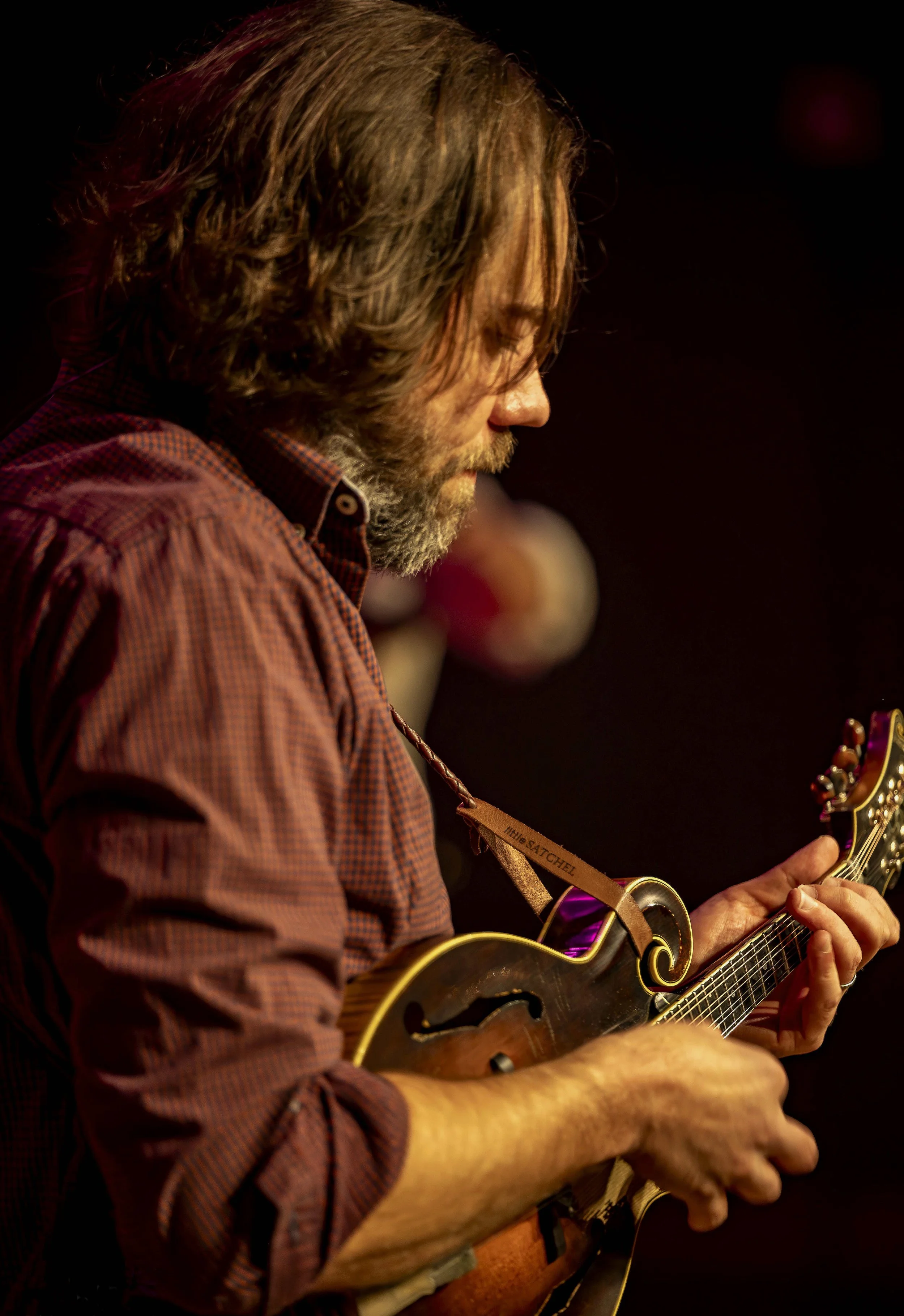 A man with long brown hair and a beard playing a vintage-style guitar against a dark background.