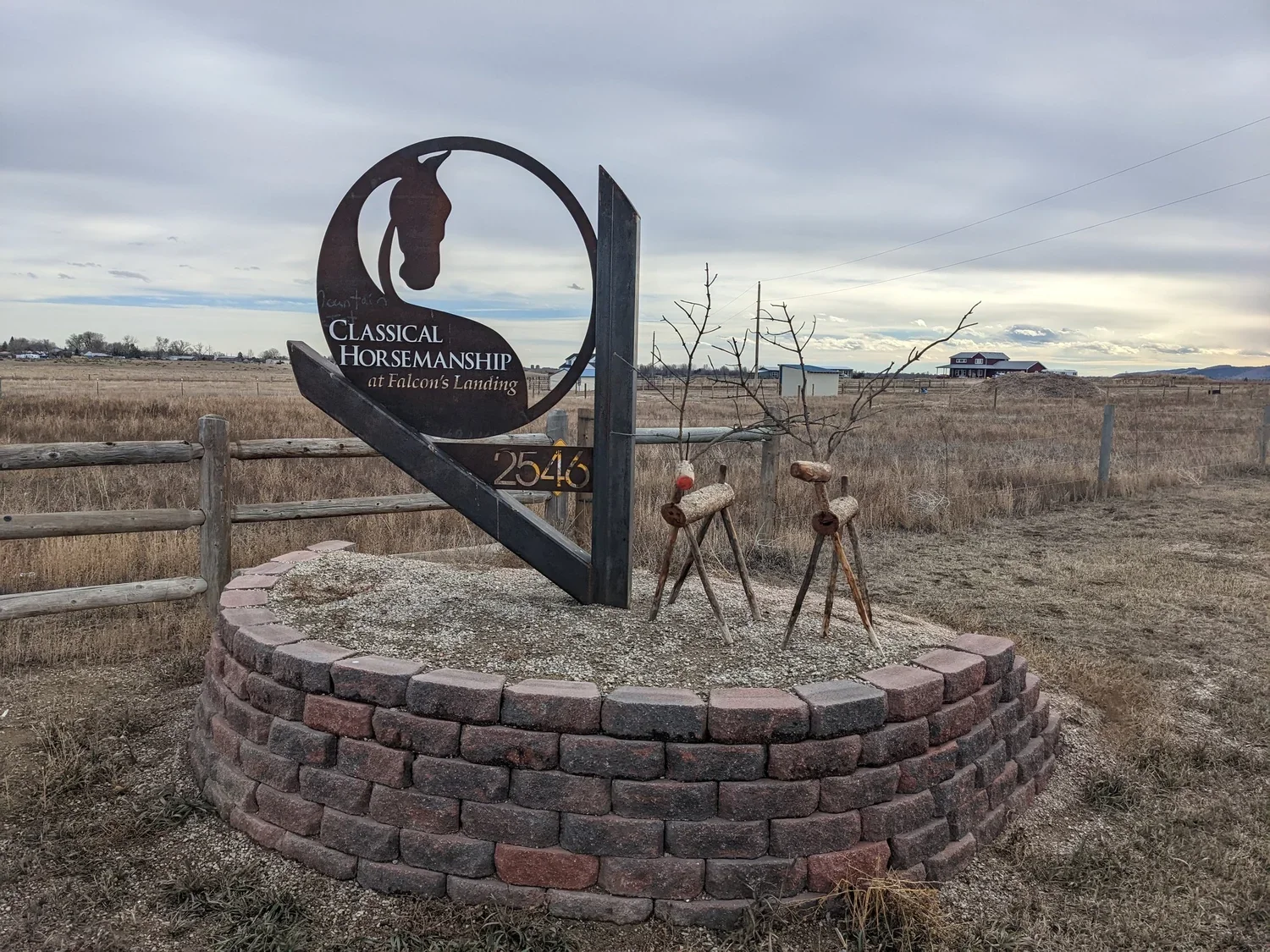 Sign for Classical Horsemanship at Falcon's Landing, with a silhouette of a horse's head inside a circle, mounted on a brick and concrete base, over an open field with a fence and a few buildings in the background under a cloudy sky.