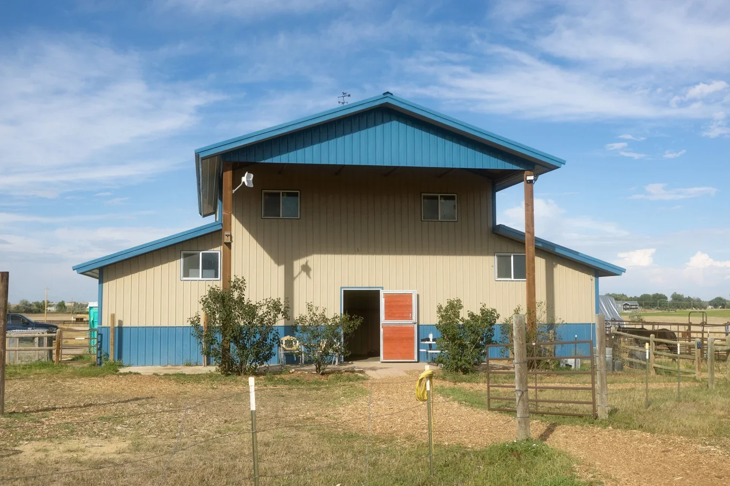 A two-story barn with beige walls, blue trim, and a sloped roof situated in a rural area. There are small bushes in front, a dirt path leading to the entrance, and a fenced pasture with horses on the right. The sky is partly cloudy.