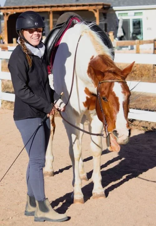 Woman in riding gear standing next to a white and brown horse with a saddle in an outdoor riding arena.