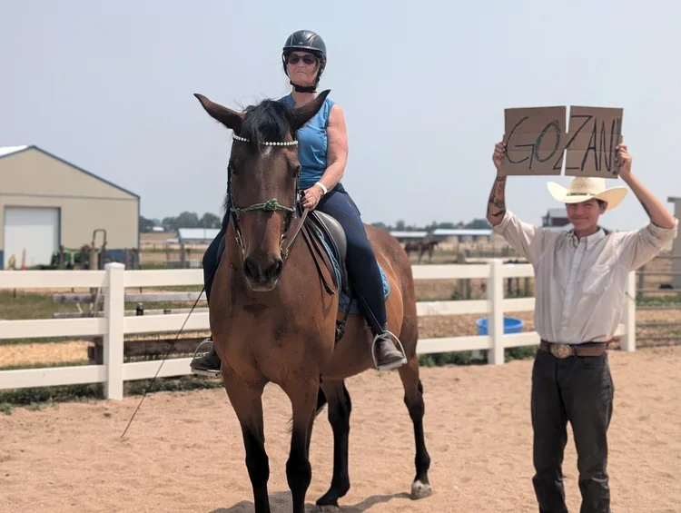 A woman in riding gear wearing a black helmet, sunglasses, and a blue vest, sitting on a brown horse in an outdoor riding arena. A young man in a white shirt, cowboy hat, and dark pants stands next to her, holding a cardboard sign that says 'COZAN'.