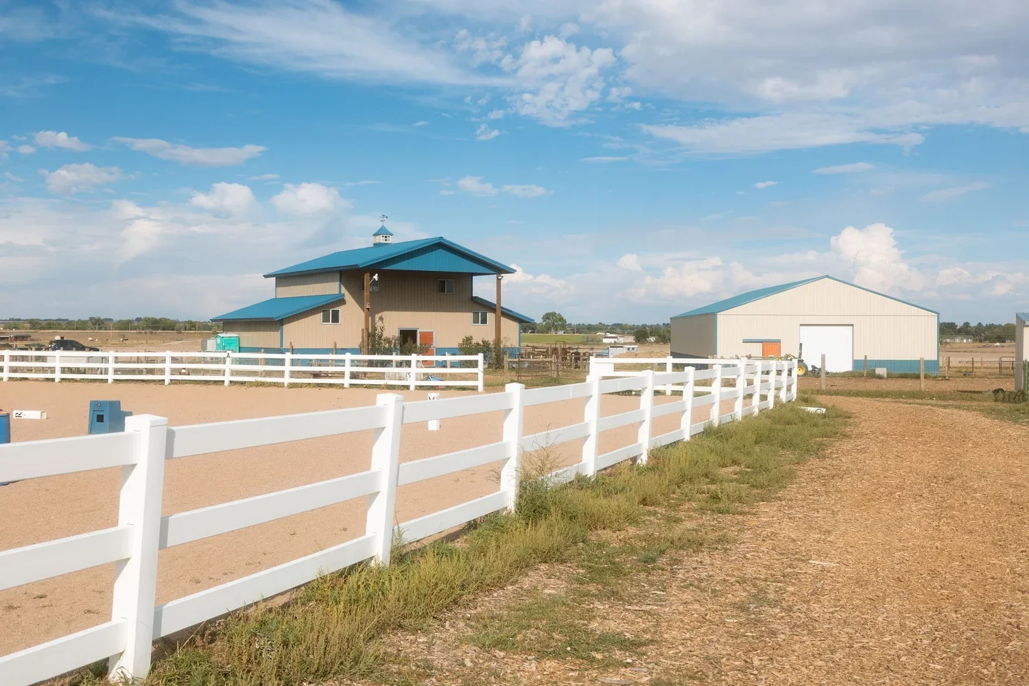 A rural farm scene with a tan two-story barn with a blue roof, a white metal shed, a gravel pathway, and white fencing under a partly cloudy sky.
