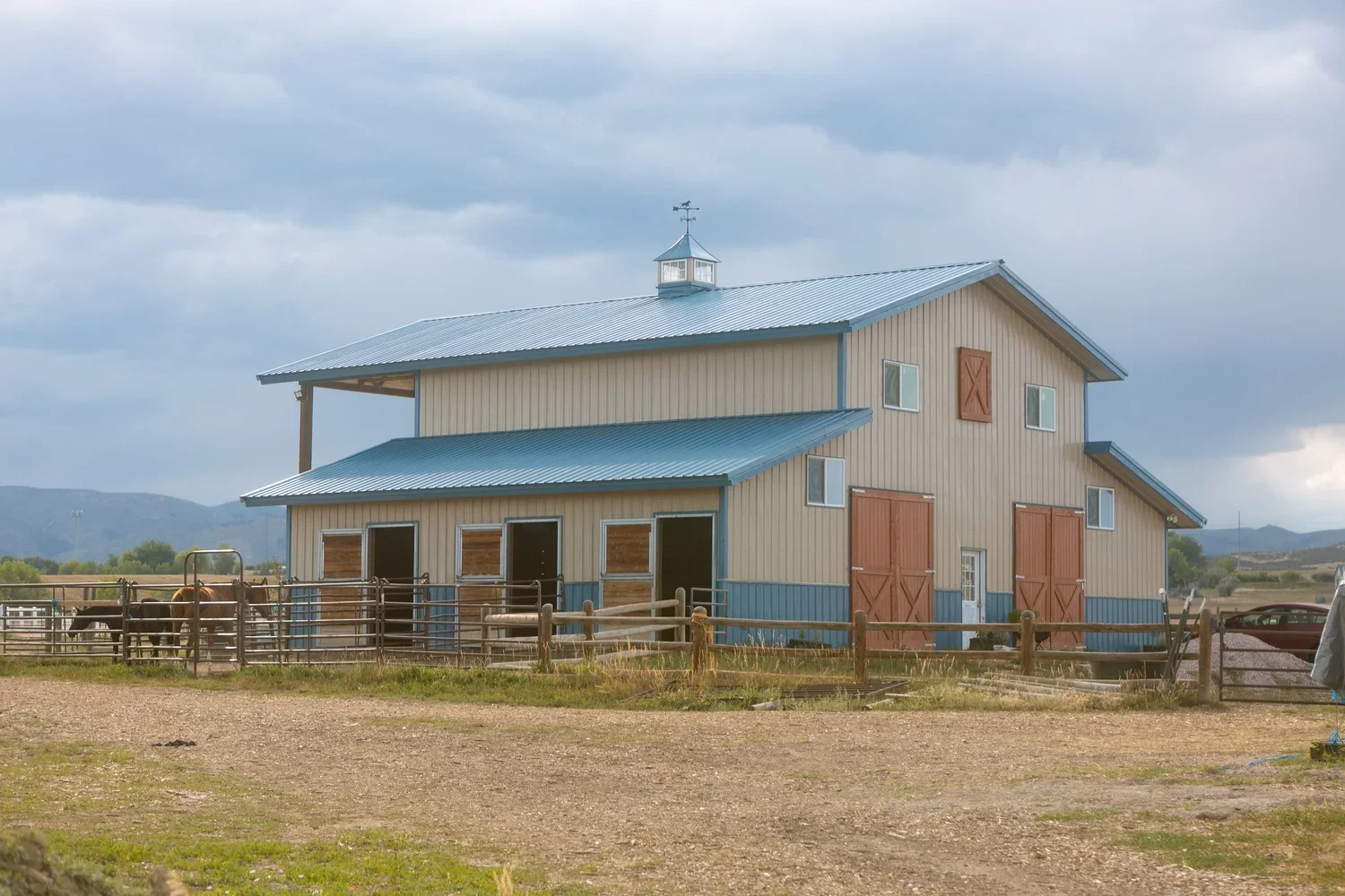 A large barn with blue metal roof and tan walls, featuring red barn doors and windows, situated in a rural landscape with a fenced area containing horses and a tractor, under cloudy skies.