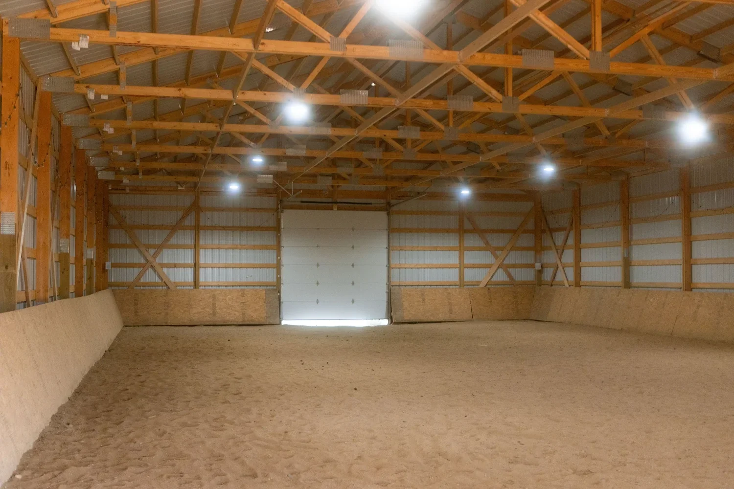 Interior of an empty indoor riding arena with a sandy floor, wooden wall panels, and metal siding on the walls and ceiling, with several ceiling lights and a large closed overhead door at the far end.