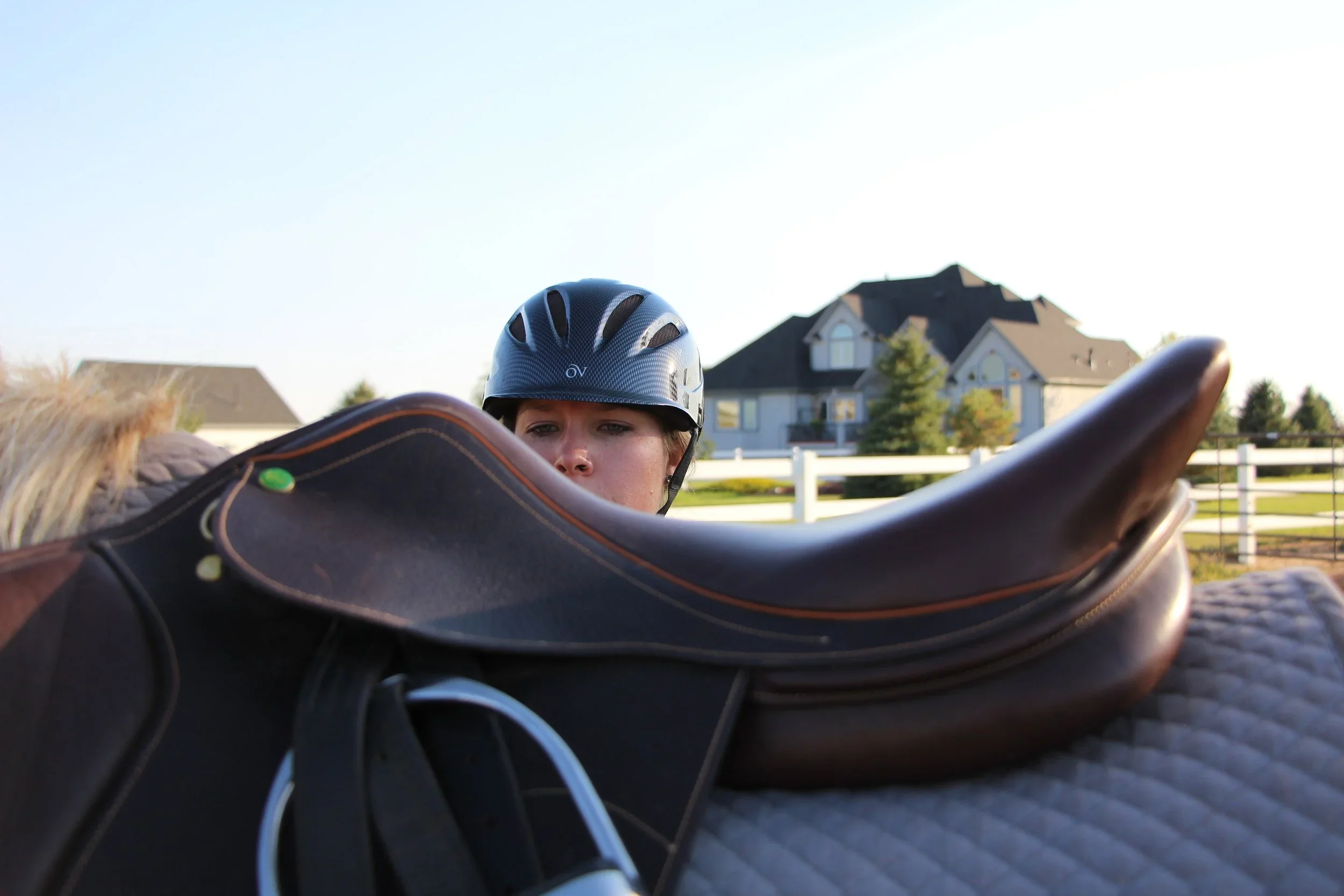 A person wearing a helmet looking over a horse's saddle on a chestnut horse in a suburban backyard.