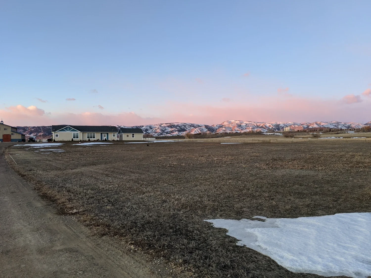 A rural landscape at sunset with a house, mountains in the background, patches of snow, and a dirt road in the foreground.