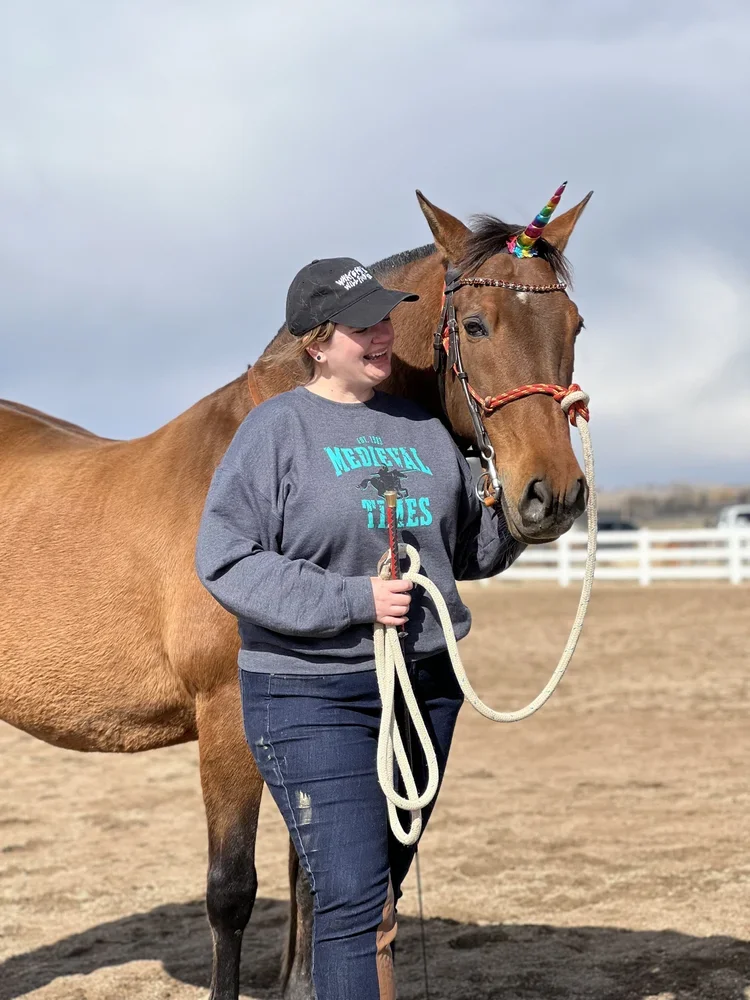 Woman with a baseball cap and jeans holding a lead rope next to a brown horse with a rainbow unicorn horn and decorative bridle at an outdoor riding arena.