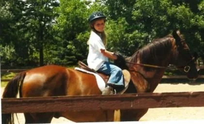 A young girl riding a horse at an outdoor riding area with trees in the background.