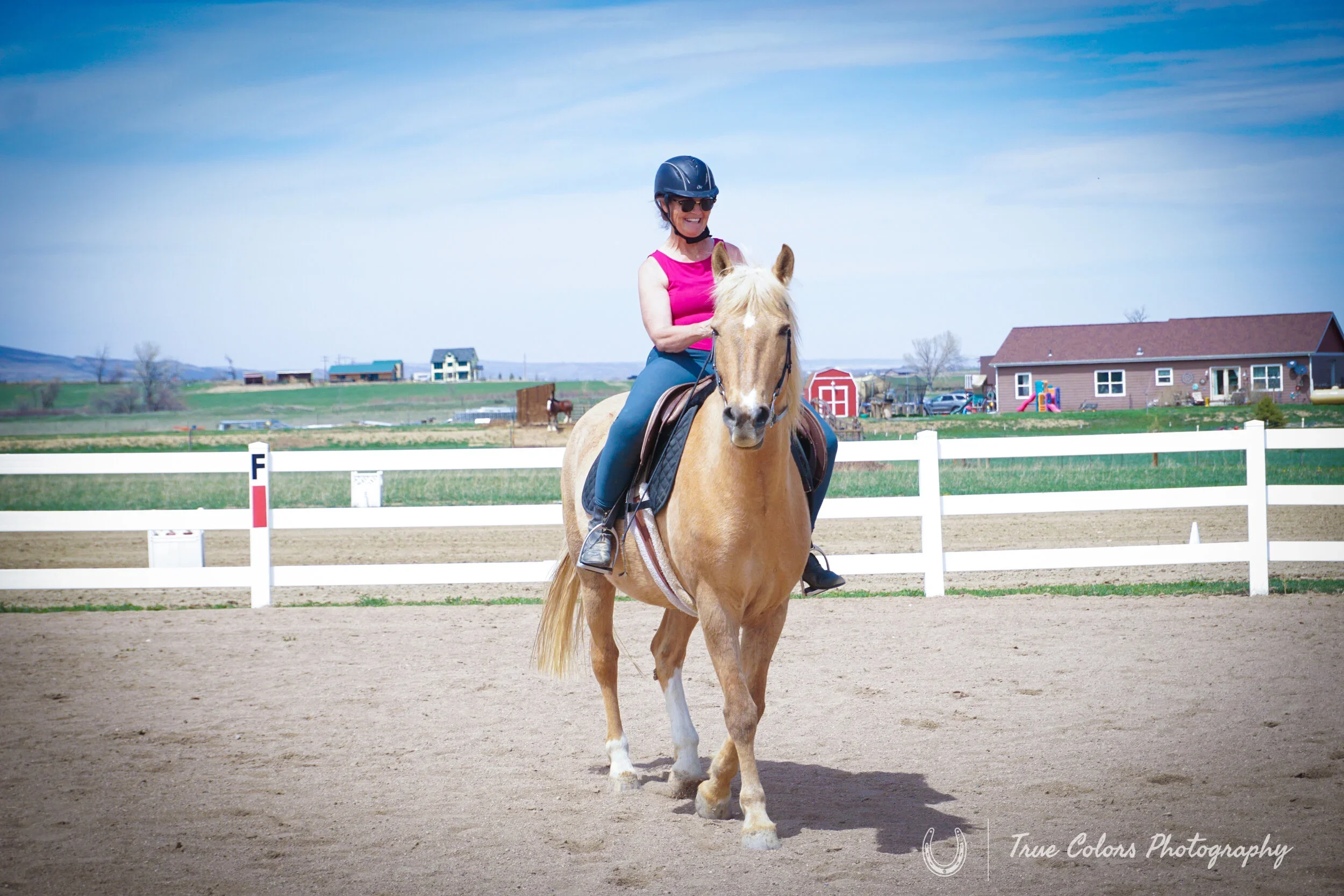 Woman riding a palomino horse on a dirt racetrack, wearing a helmet and pink top, with farm buildings and blue sky in the background.