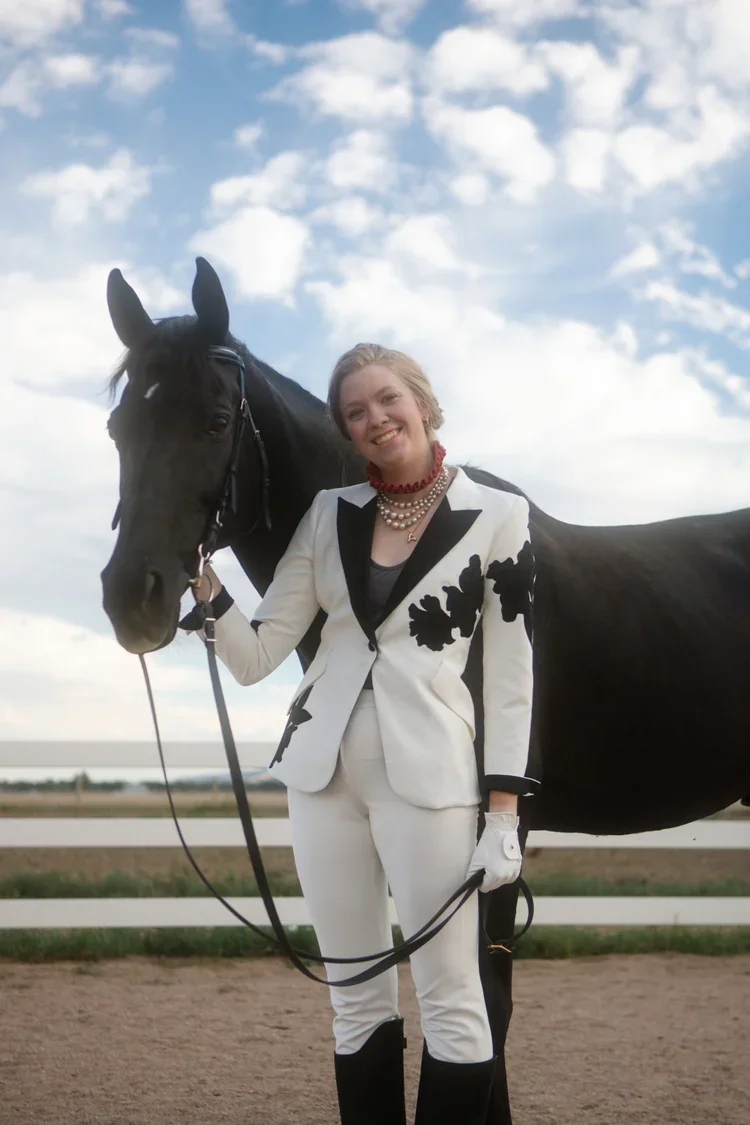 Woman in a white suit with black floral patterns holding a black horse during daytime.