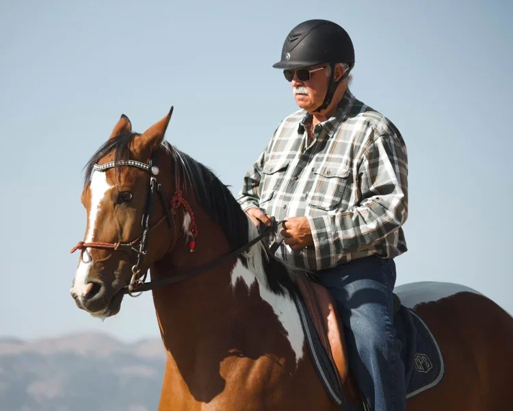 An older man wearing a plaid shirt, helmet, and sunglasses riding a brown and white horse under a clear sky.
