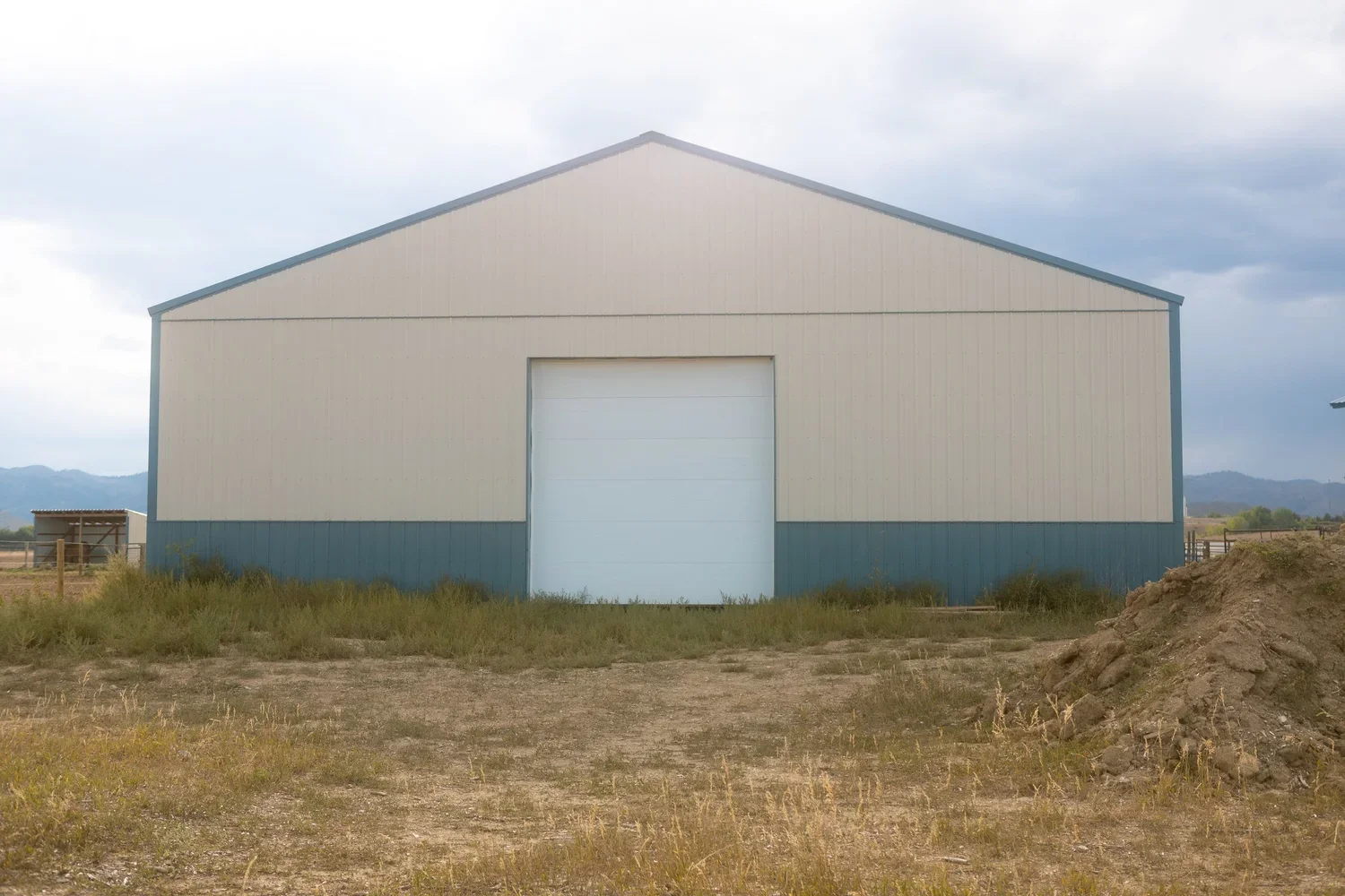 Large beige and blue metal barn with a closed white garage door, situated in a rural area with dirt and grass, hills in the distance, and cloudy sky overhead.