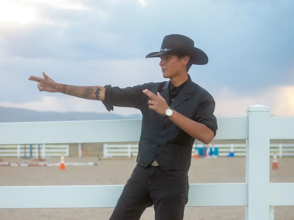 Person dressed as a cowboy with a black hat and black outfit, standing outdoors near a white fence, with cloudy sky in the background, striking a pose with one arm extended and fingers pointed.