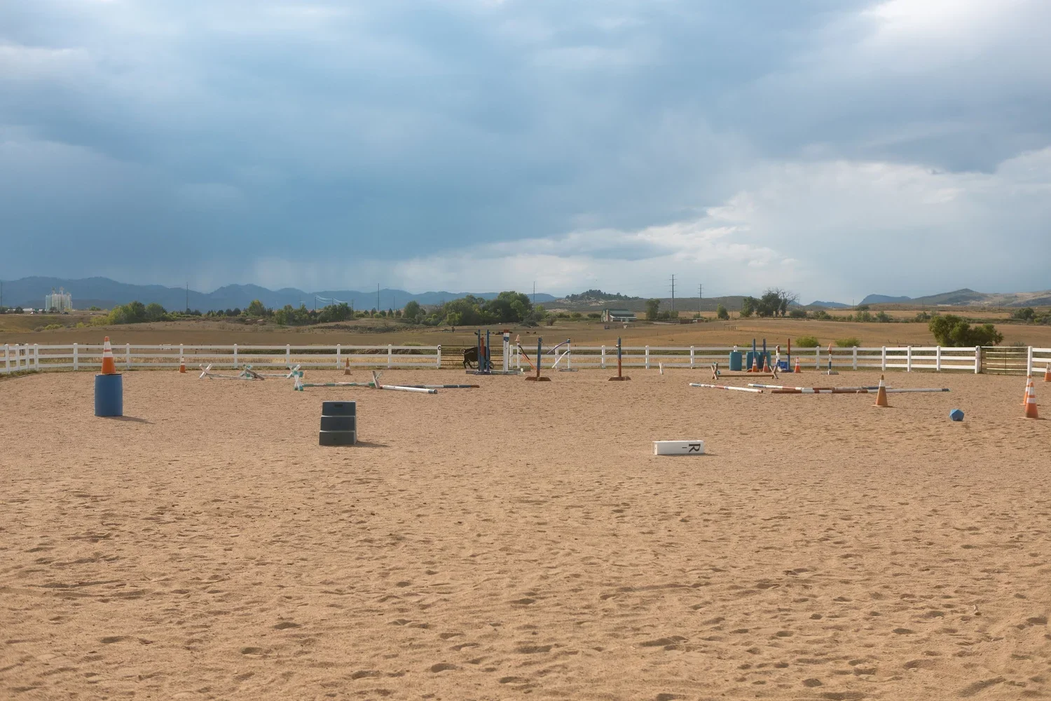 An outdoor horse jumping arena with jump obstacles, traffic cones, and barriers, surrounded by a white fence, with a scenic landscape of mountains and cloudy sky in the background.