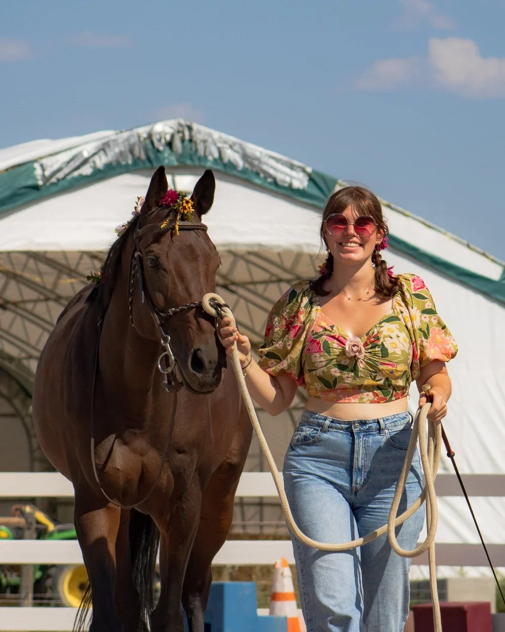 A young woman with brown hair in pigtails, wearing pink sunglasses, a floral crop top, and jeans, holding a brown horse by the reins, outdoors on a sunny day with a tent and construction cones in the background.
