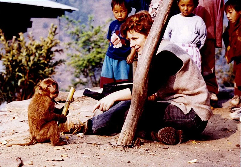 A woman interacting with a monkey holding a small banana in an outdoor setting, with children observing nearby.