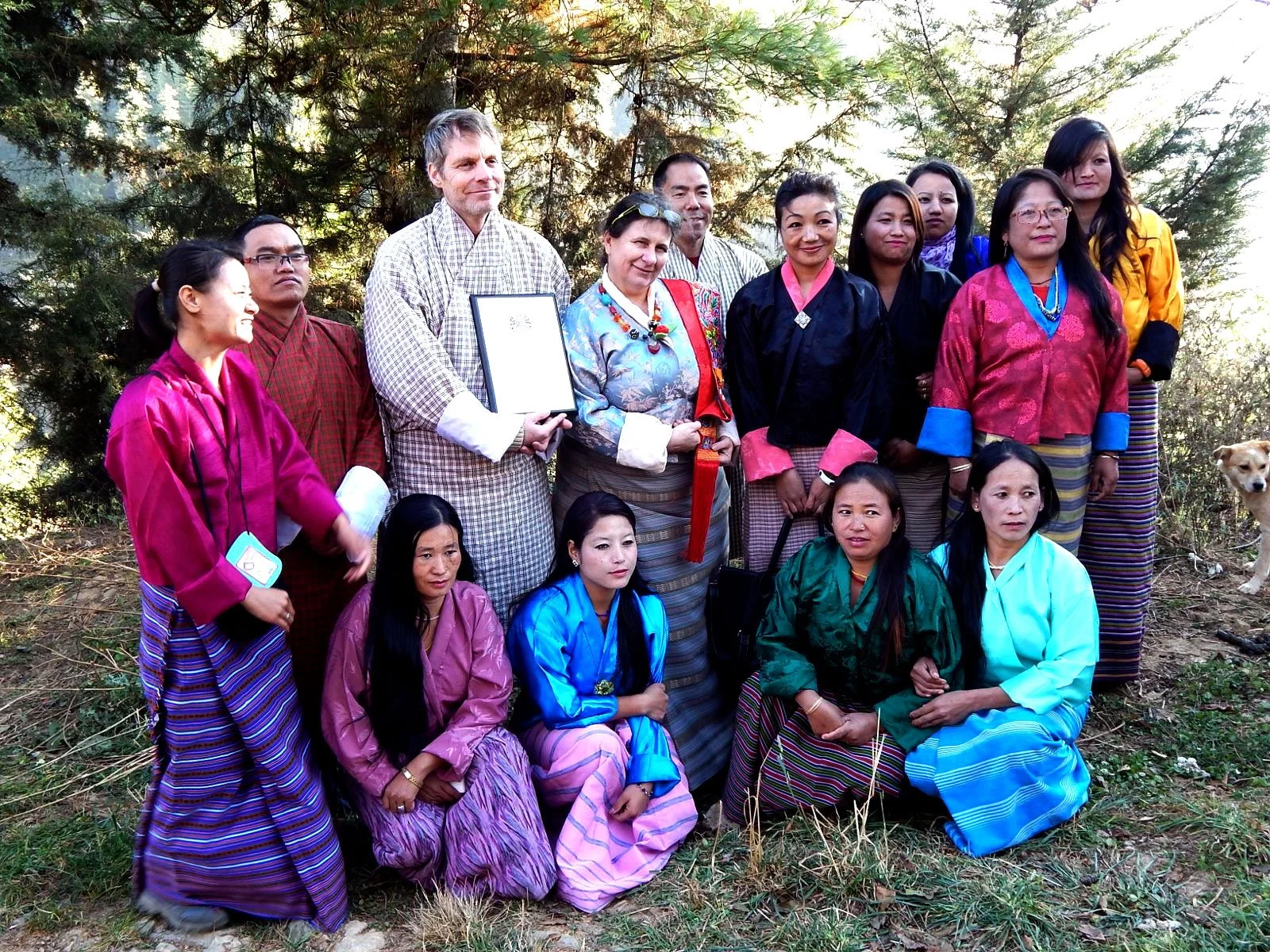 Group of people dressed in traditional Bhutanese clothing outdoors, with trees in the background, some individuals kneeling in front and others standing behind, including a dog on the right.