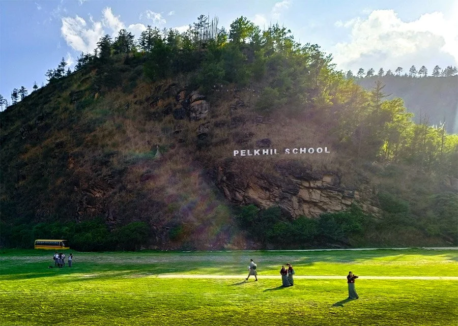 A hillside with trees and rocks, with the words 'Pelkih School' on the hillside, a grassy field in the foreground where children are playing or walking, and a school bus parked near some bushes.