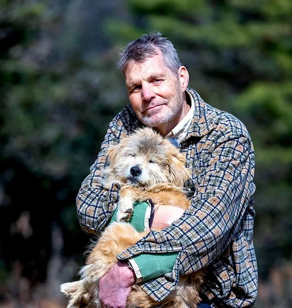 A man with gray hair and a beard holding a fluffy puppy outdoors with a blurred green background.