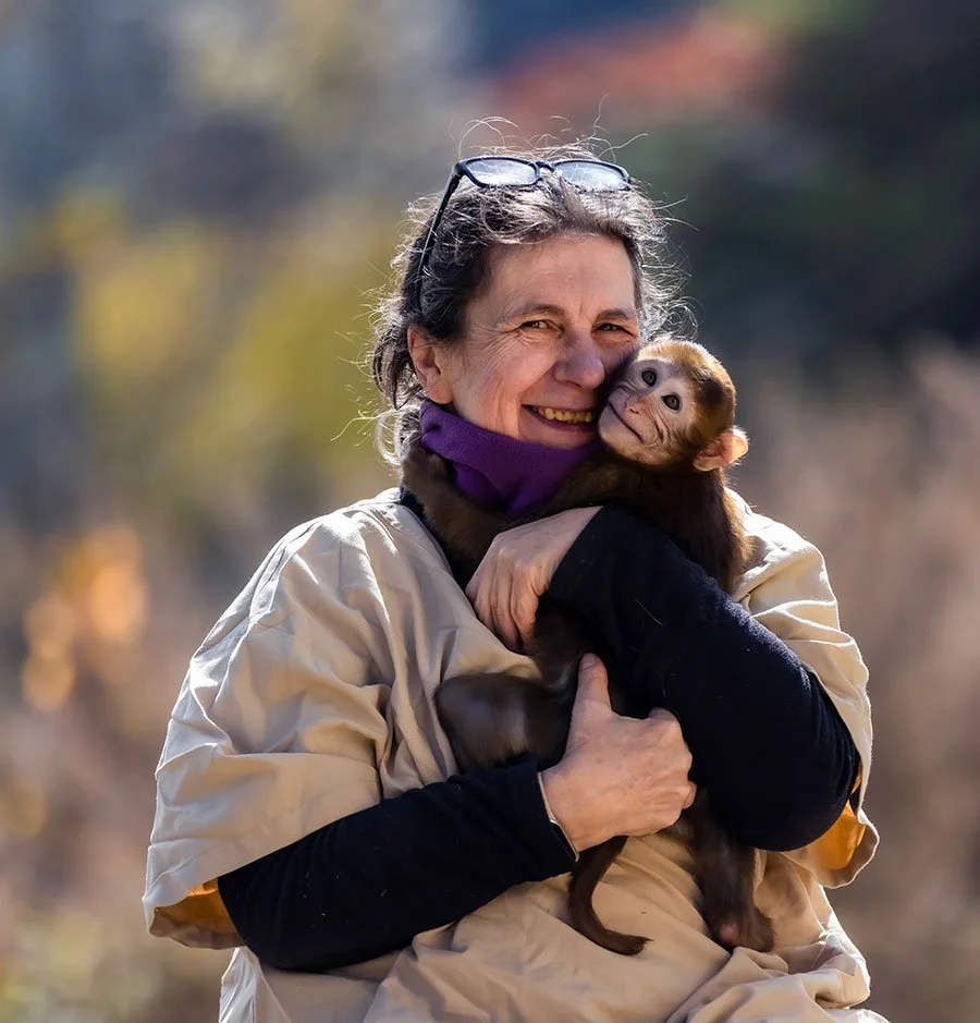 A woman smiling and hugging a baby monkey outdoors during daytime, with trees and autumn leaves in the background.