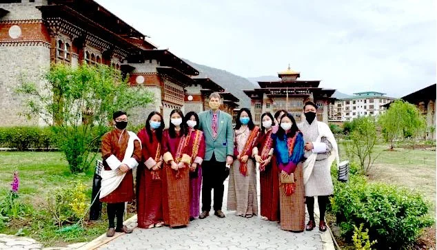Group of seven people standing outdoors in front of traditional Bhutanese buildings, with mountains in the background, all wearing face masks and traditional attire.