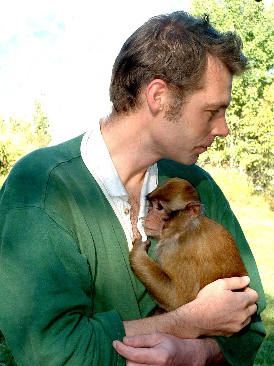 A man with brown hair and a green jacket holding a small monkey close to his chest outdoors, with trees and a blue sky in the background.