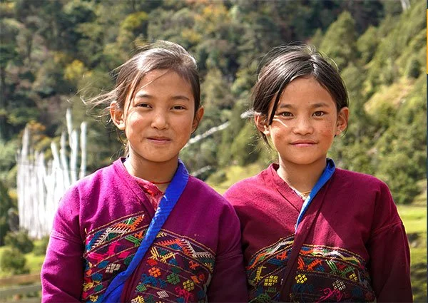 Two young girls wearing colorful traditional clothing outdoors with greenery and a waterfall in the background.