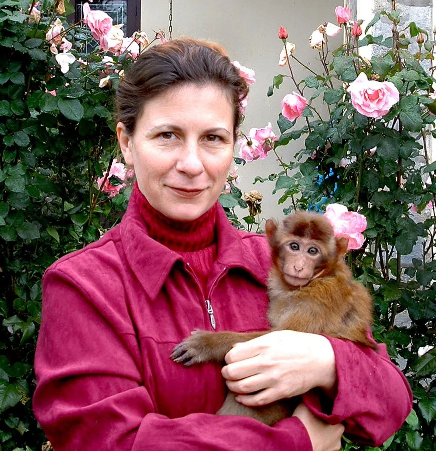 A woman with brown hair in a red coat holding a baby monkey with brown fur, in front of pink roses.