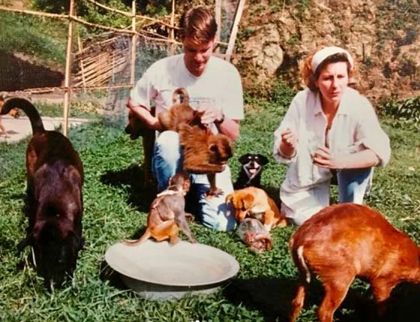 Two women surrounded by various puppies and dogs outdoors on green grass.