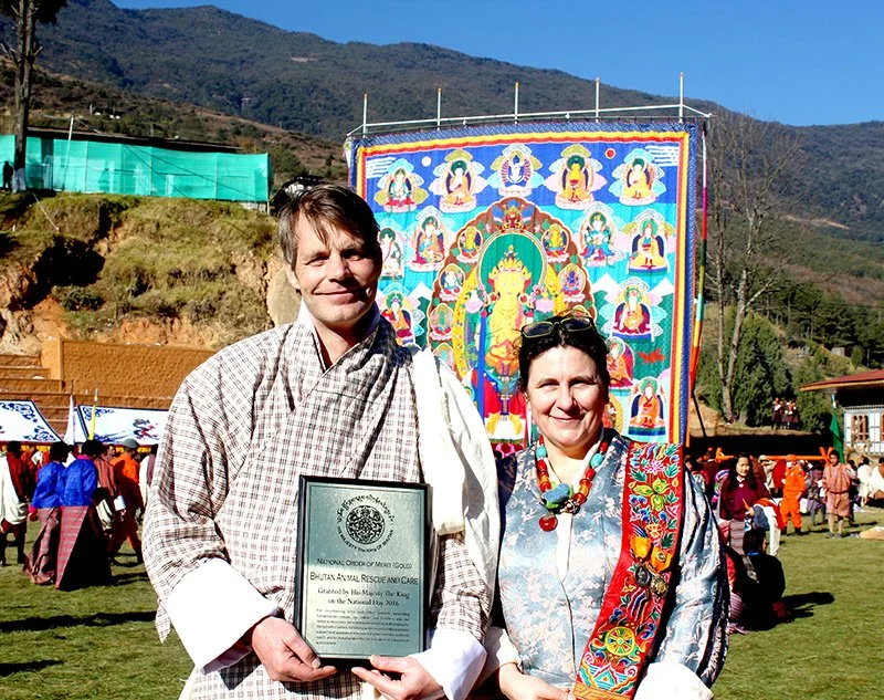 Two people standing outdoors, smiling. The person on the left is dressed in traditional Bhutanese attire and holding a plaque. The person on the right wears colorful traditional clothing with a red embroidered sash. Behind them, a large colorful tapestry or thangka featuring a Buddhist deity or figure is displayed. There are other people in traditional clothing in the background, with mountains and clear blue sky overhead.