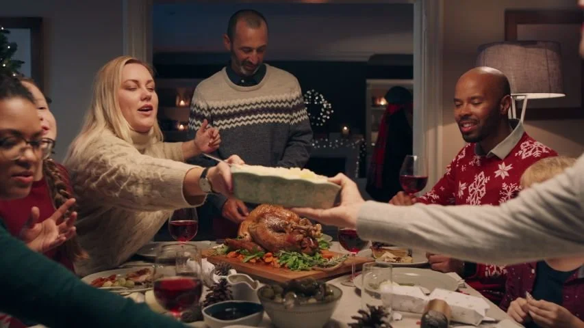 Family gathered around a decorated dining table sharing a holiday meal, serving turkey, with wine glasses and festive decorations in the background.