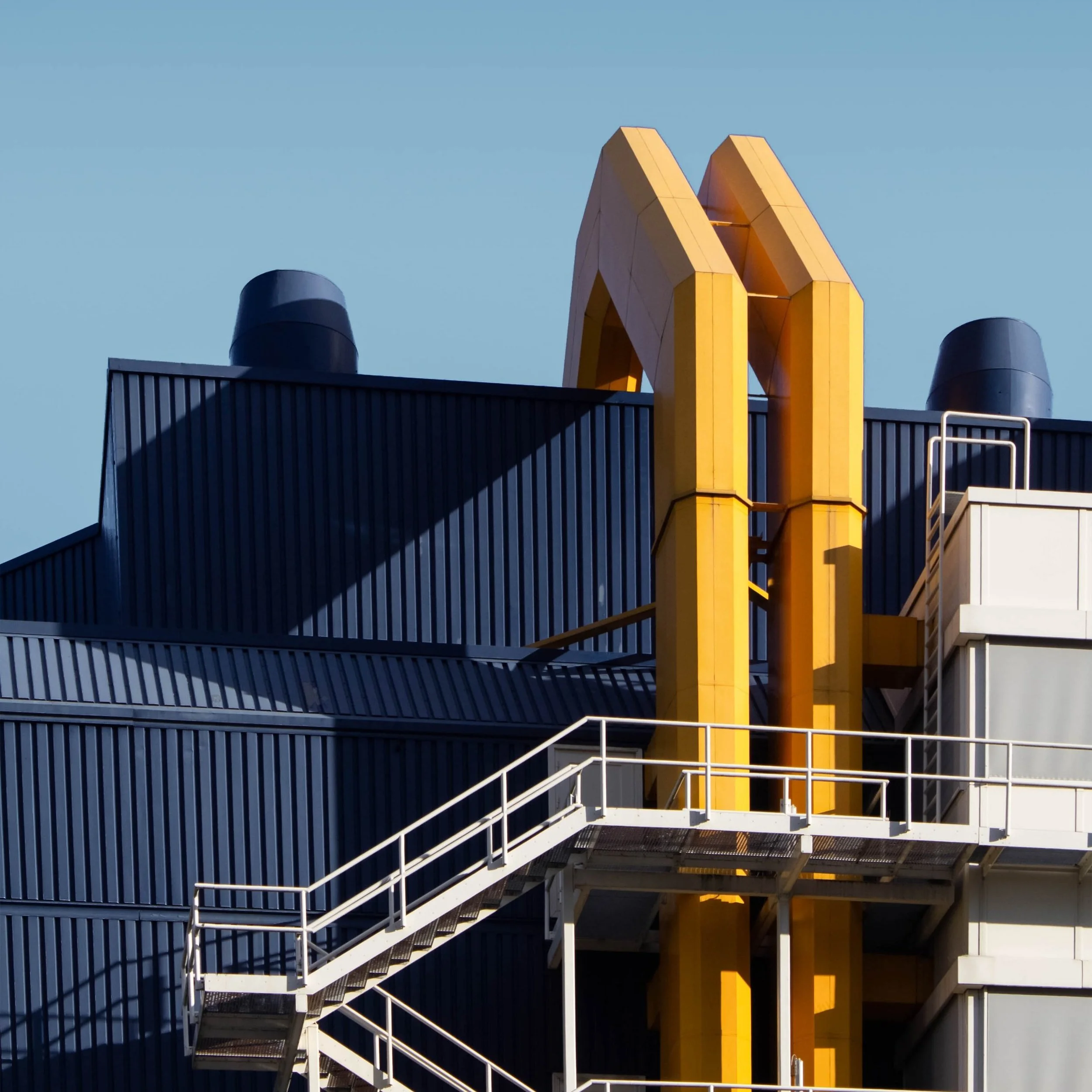 Close-up of a modern industrial building with dark blue corrugated metal walls, white fire escape stairs, and yellow ventilation pipes against a clear blue sky.
