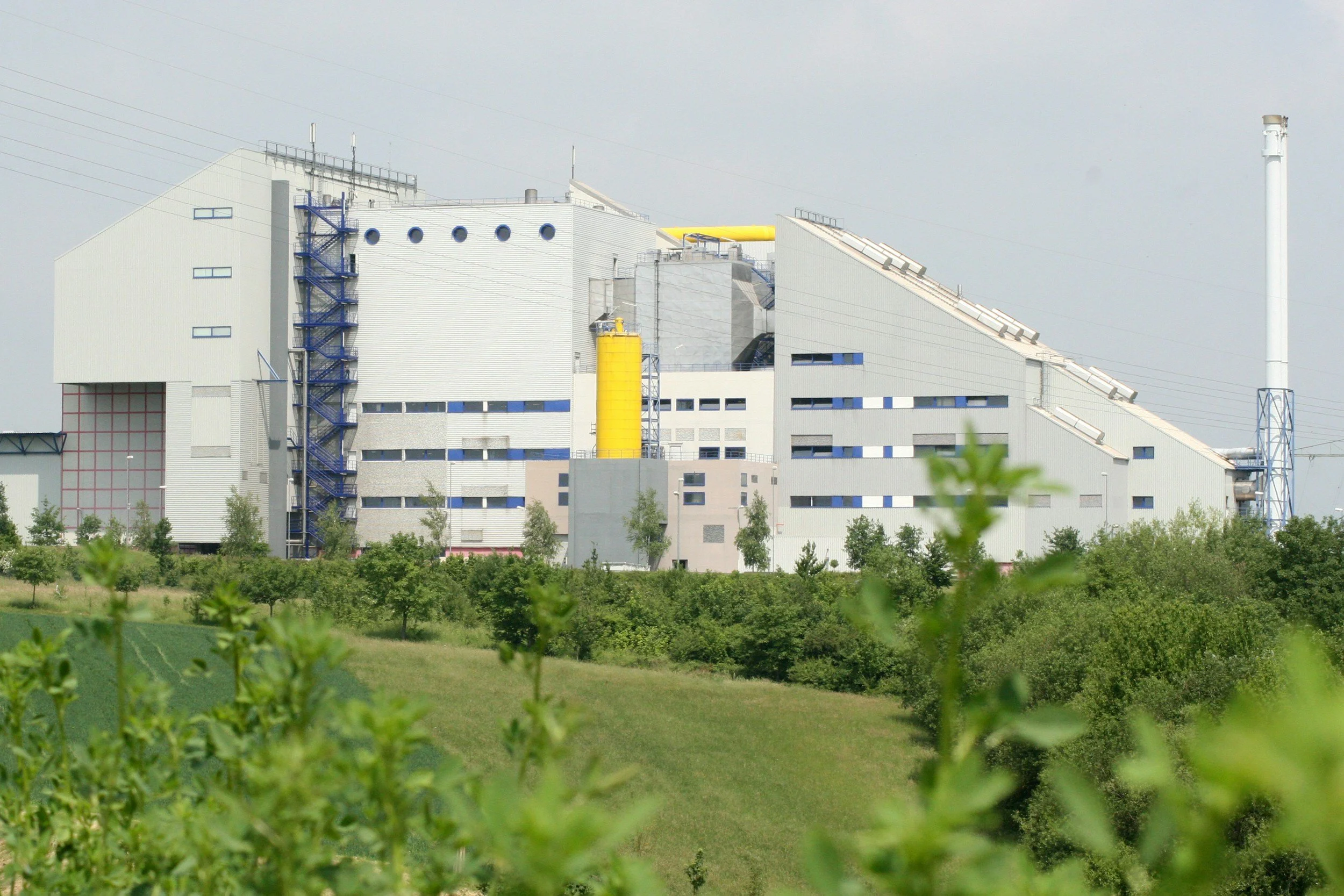 Large industrial building or power plant with blue and white exterior, yellow components, and a tall smokestack, surrounded by green trees and grassy fields.