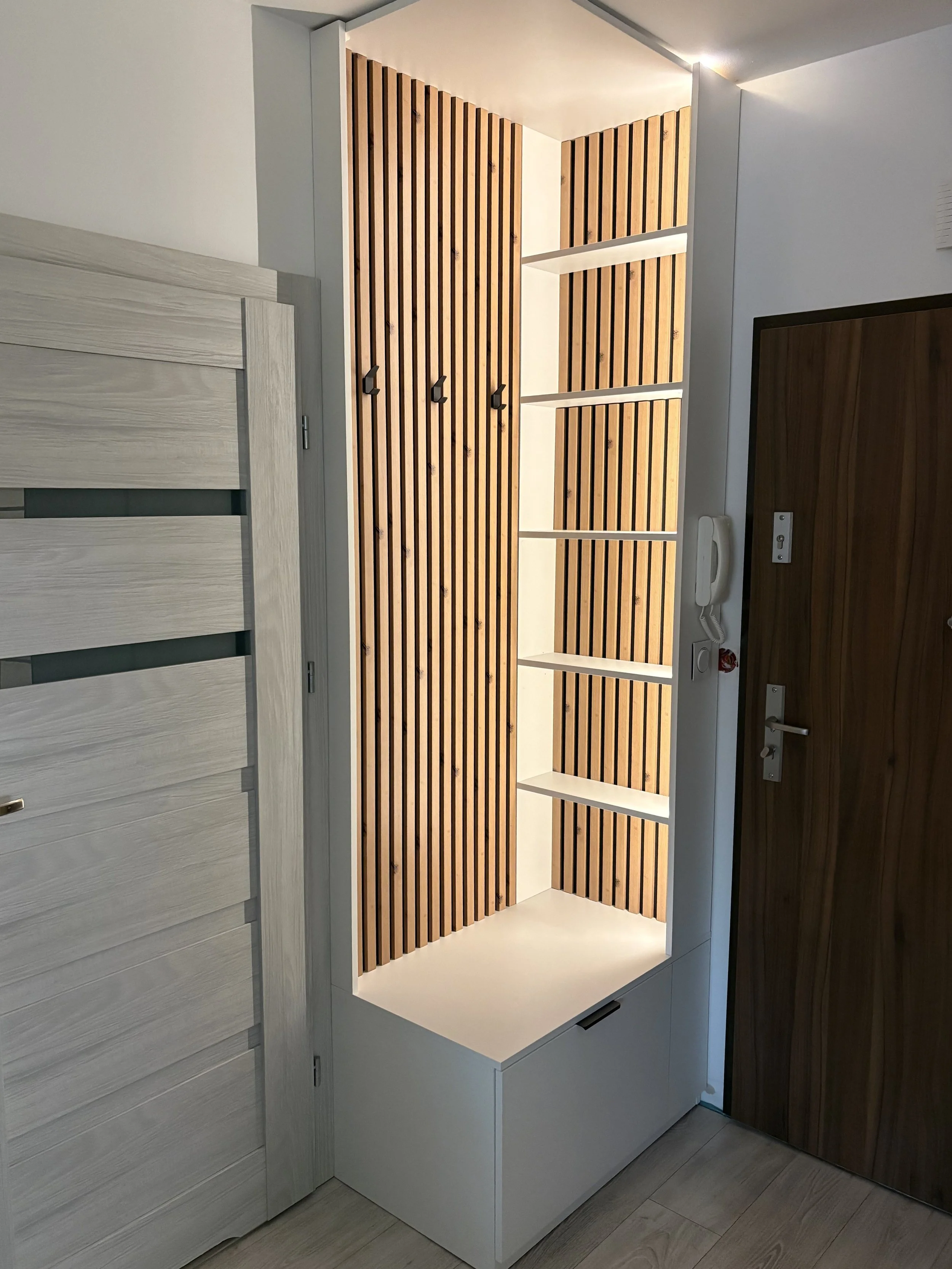 Modern hallway storage unit with wooden slats, white shelves, and hooks, positioned next to a light wooden door.