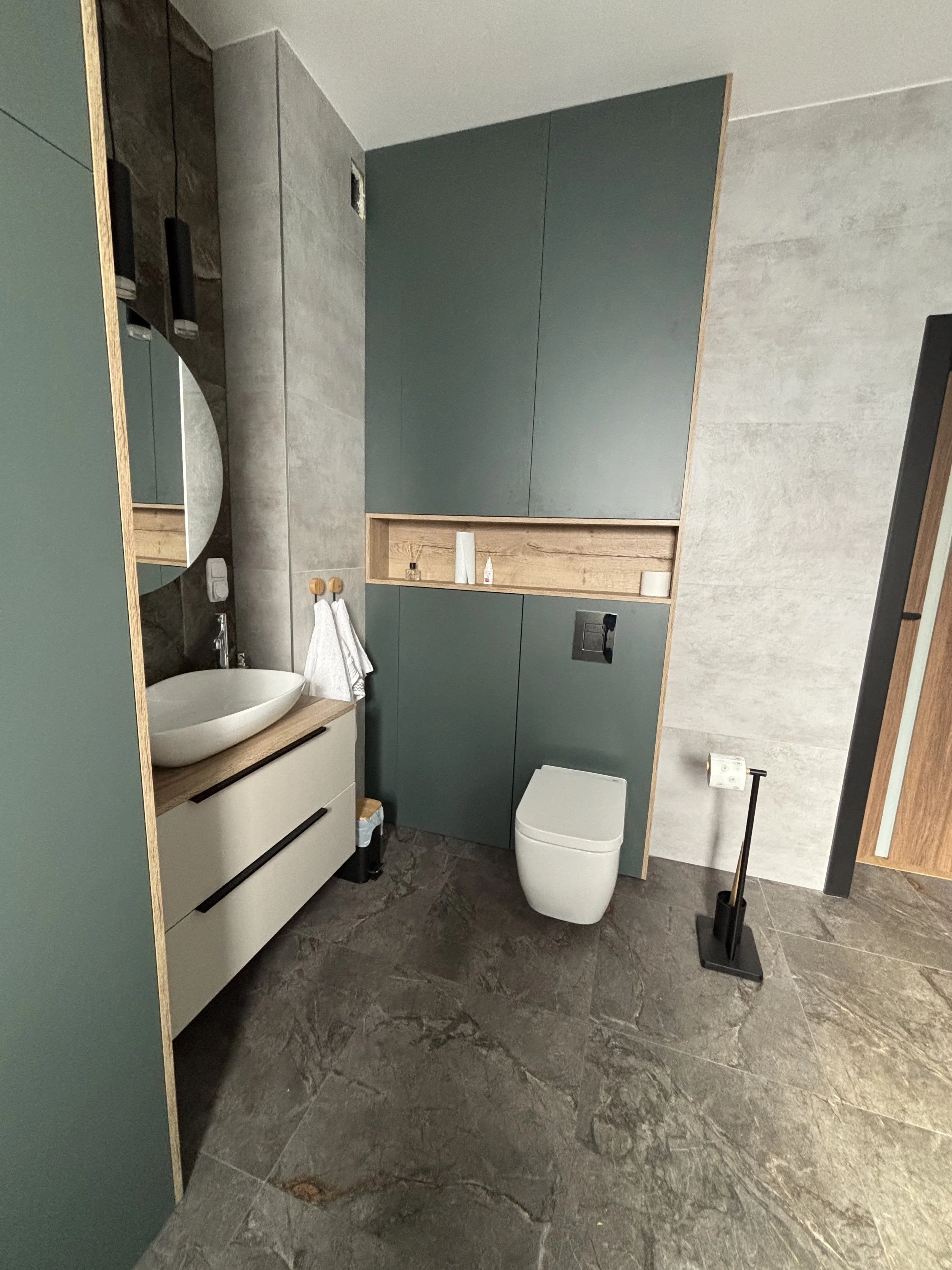 A modern bathroom with a light-colored vanity and a vessel sink on a wooden countertop. A round mirror is mounted above the sink. There is a toilet with a built-in flush system in front of a wall with concealed storage cabinets, some with open shelve