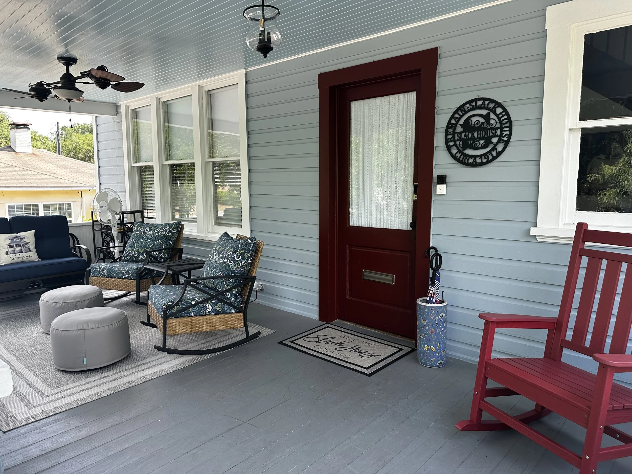 Porch area with chairs, ottomans, a ceiling fan, and a decorative sign on the wall that reads "Welcome to the Stack House Circa 1912."
