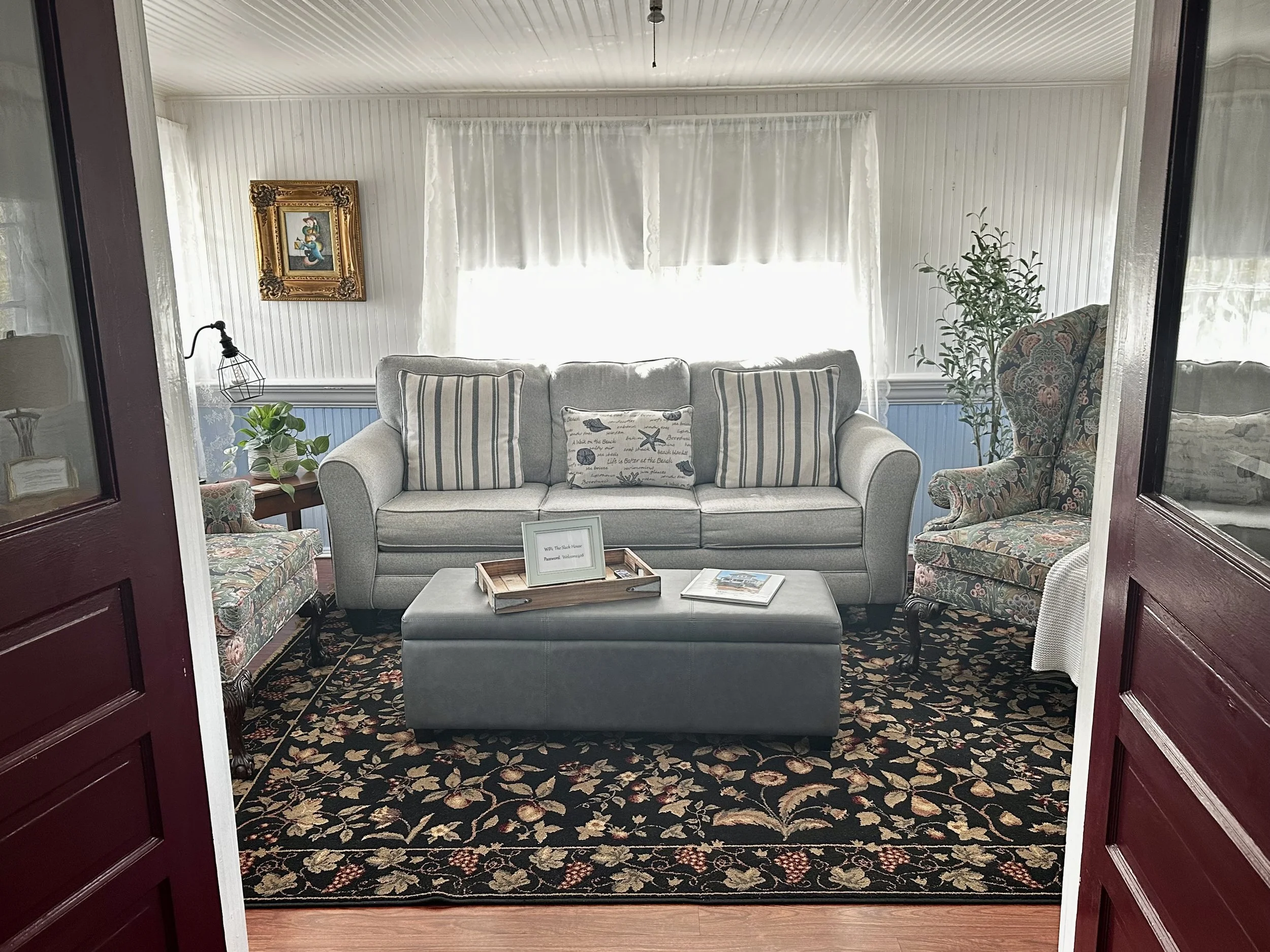 Living room seen through an open doorway with red trim. There's a light gray sofa with striped pillows in front of large windows with white curtains. To the right is a floral patterned armchair and a potted plant. A wooden table has a framed sign and a magazine. The room has a patterned rug under the furniture.