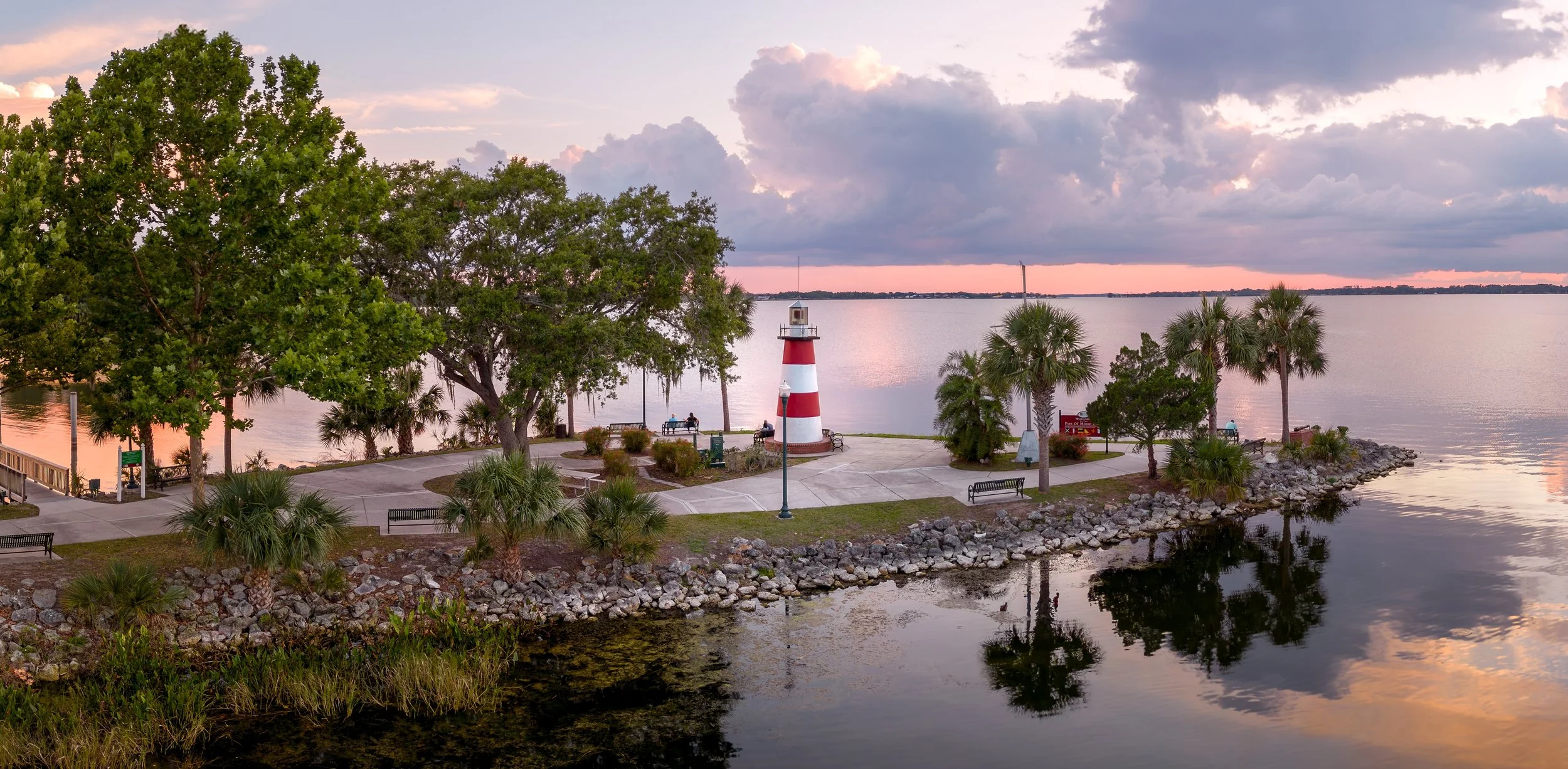 A lakeside park at sunset with a red and white lighthouse, trees, benches, and a calm body of water reflecting the sky and landscape.