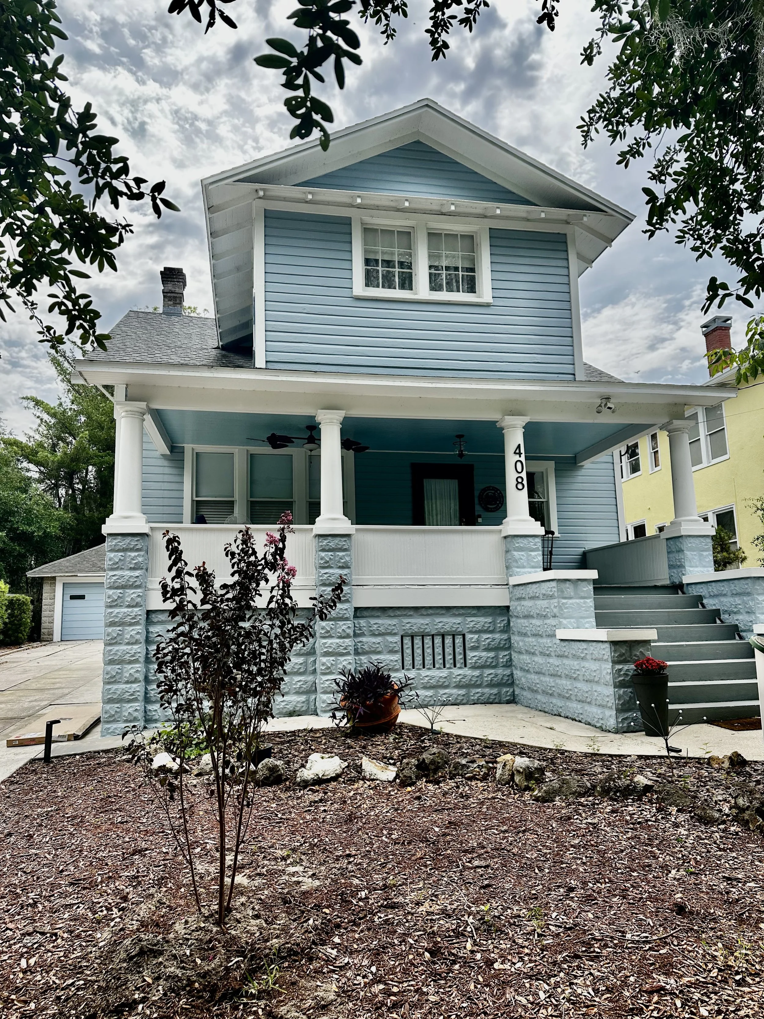 A two-story house painted in blue with white trim. It has a front porch with white columns, a staircase, and a small garden in front with plants and mulch. The sky above is cloudy with some sunlight.