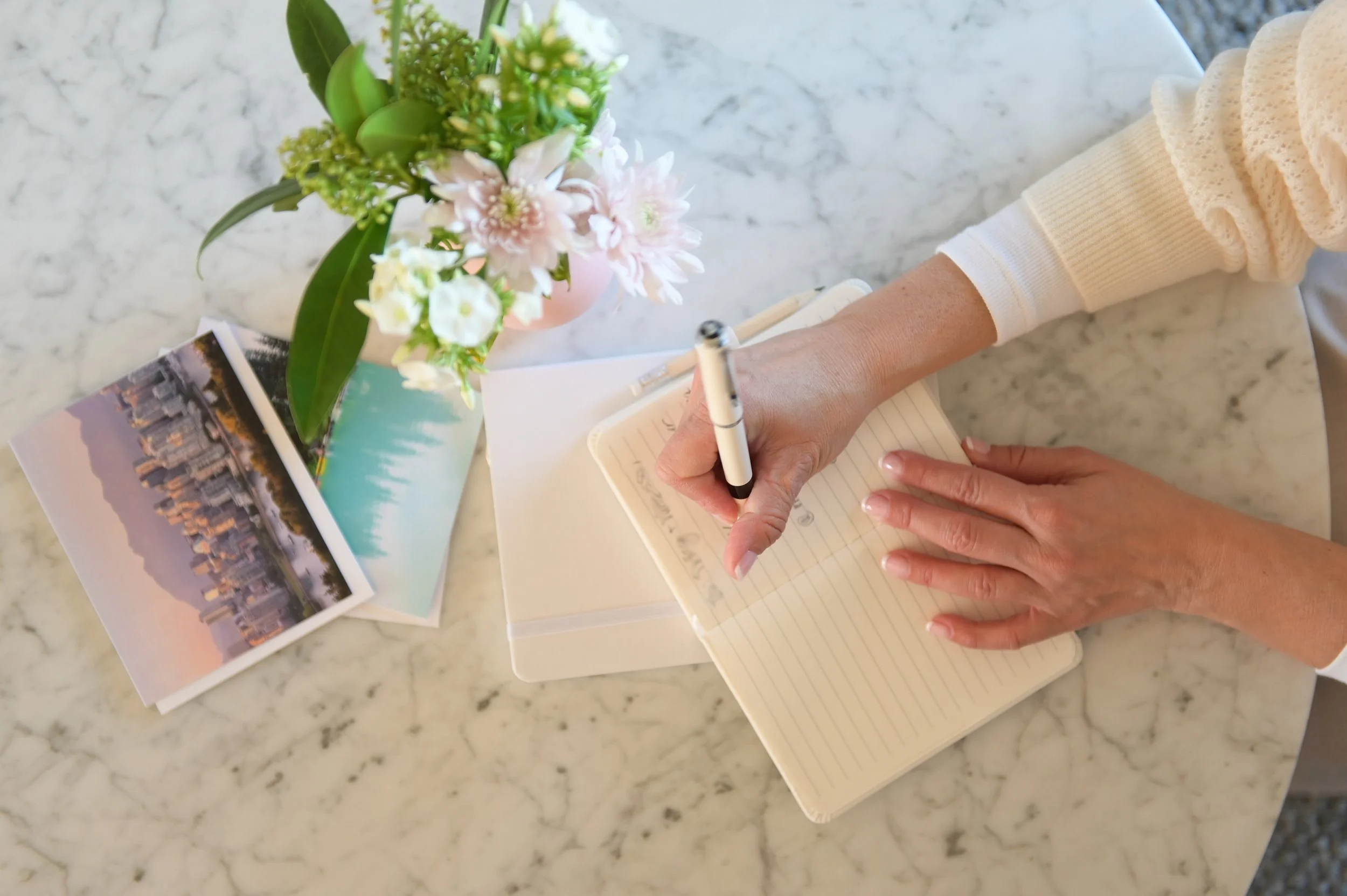 A person writing in a notebook on a marble table with a vase of pink and white flowers, postcards, and a pen.