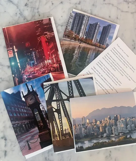 Several postcards and a printed page spread out on a marble surface. The postcards feature urban cityscapes, including a skyline with skyscrapers along a river, a street with neon lights, a clock tower, and a bridge at sunset.