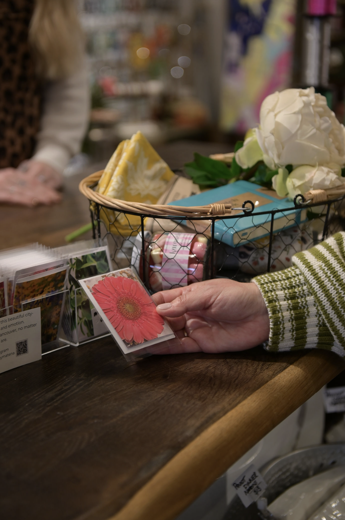 Person holding a small card with a pink flower photo at a store counter with gardening cards, flowers, and a basket of items on the counter.