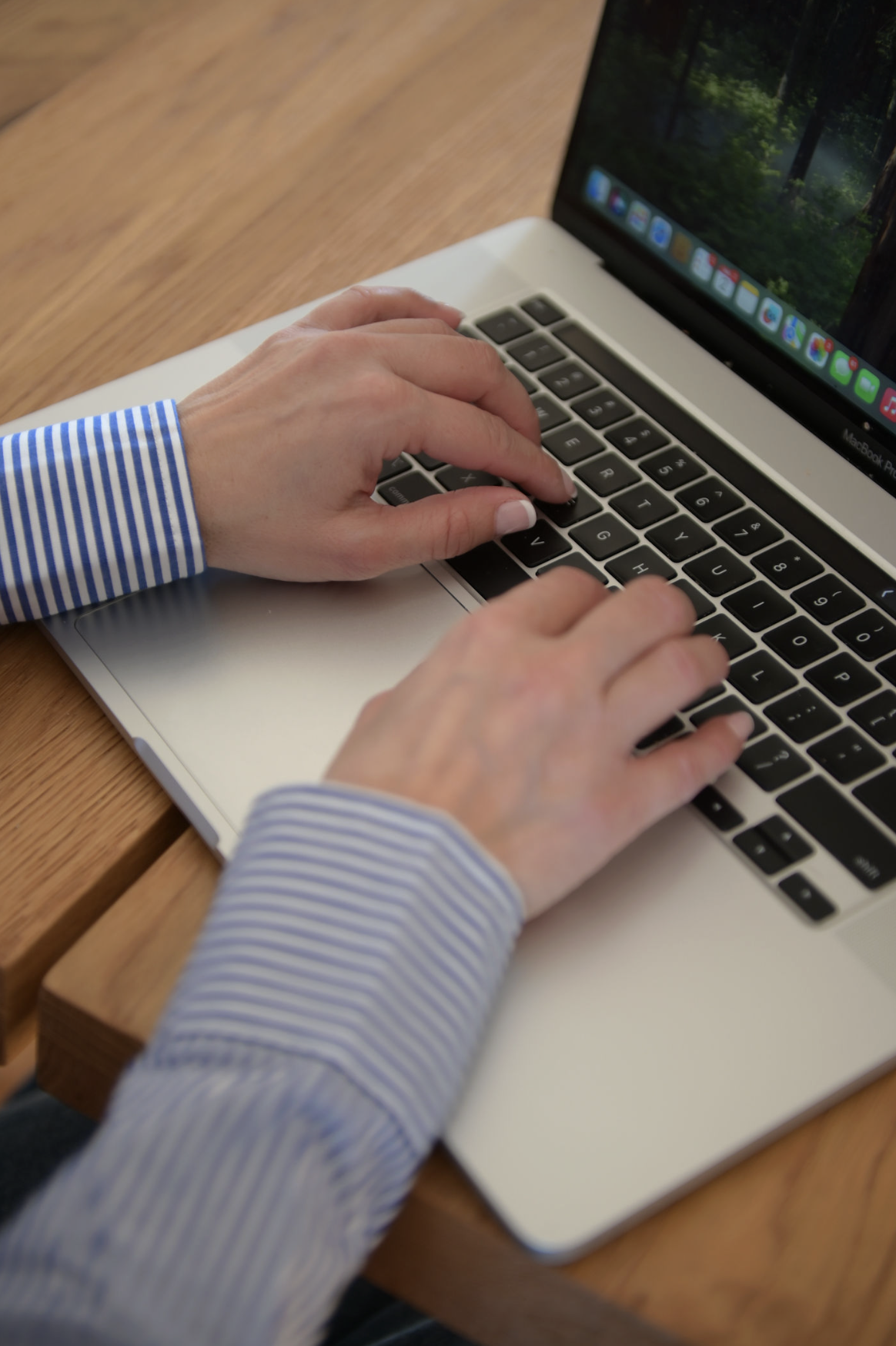 Person typing on a MacBook laptop with a nature scene wallpaper, sitting at a wooden table.