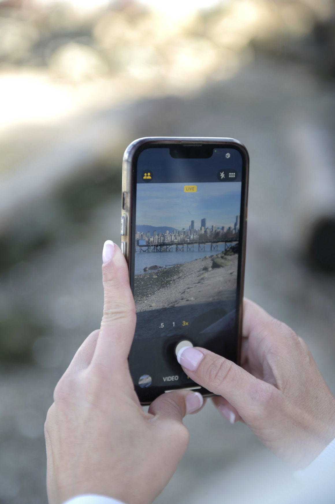 Close-up of a person holding a smartphone, capturing a city skyline with tall buildings and a bridge over water, with a blurred natural landscape in the background.