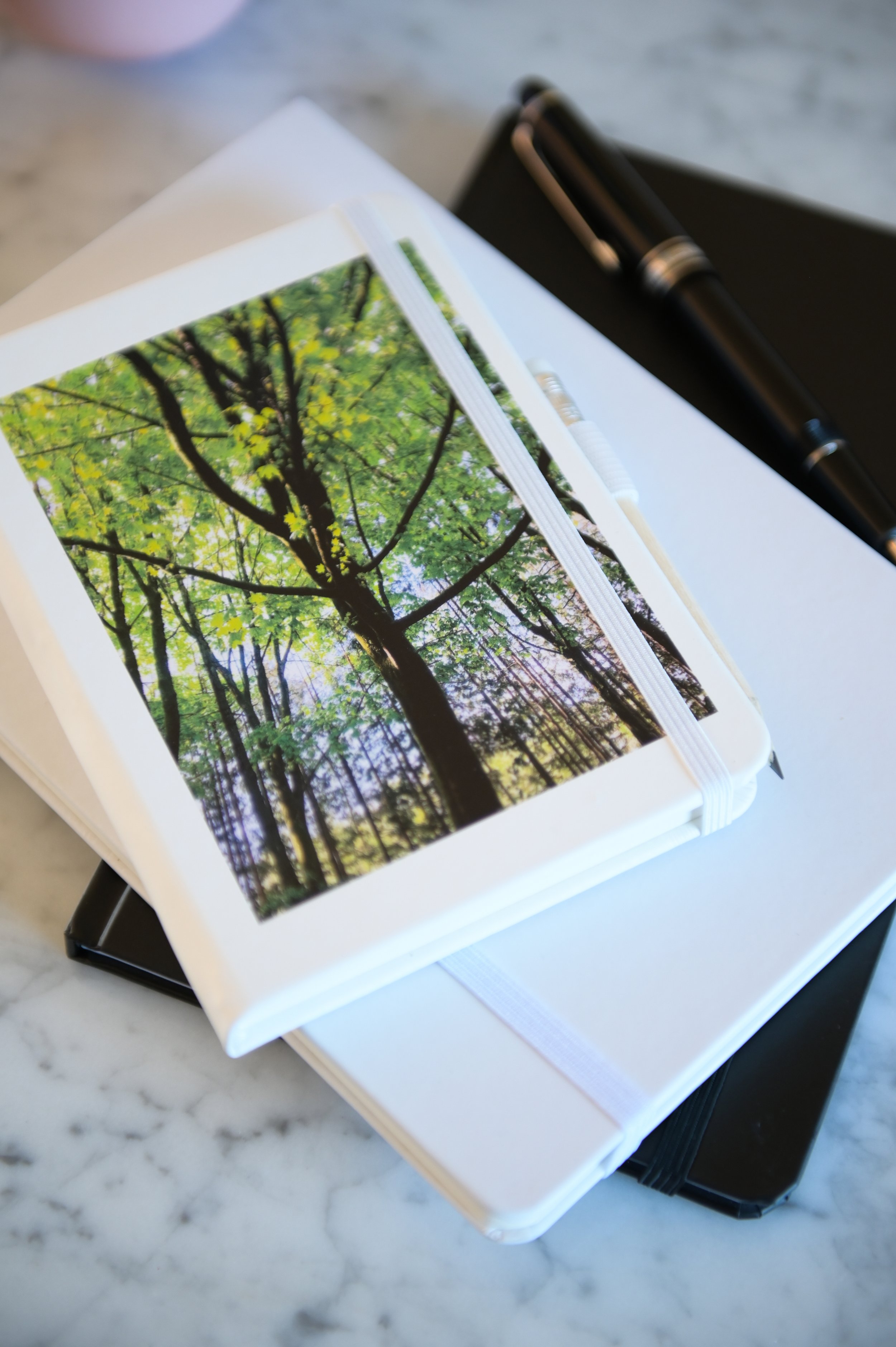 A closed hardcover book with a picture of green trees on the cover, resting on a white surface with a black binder clip and a black object nearby.