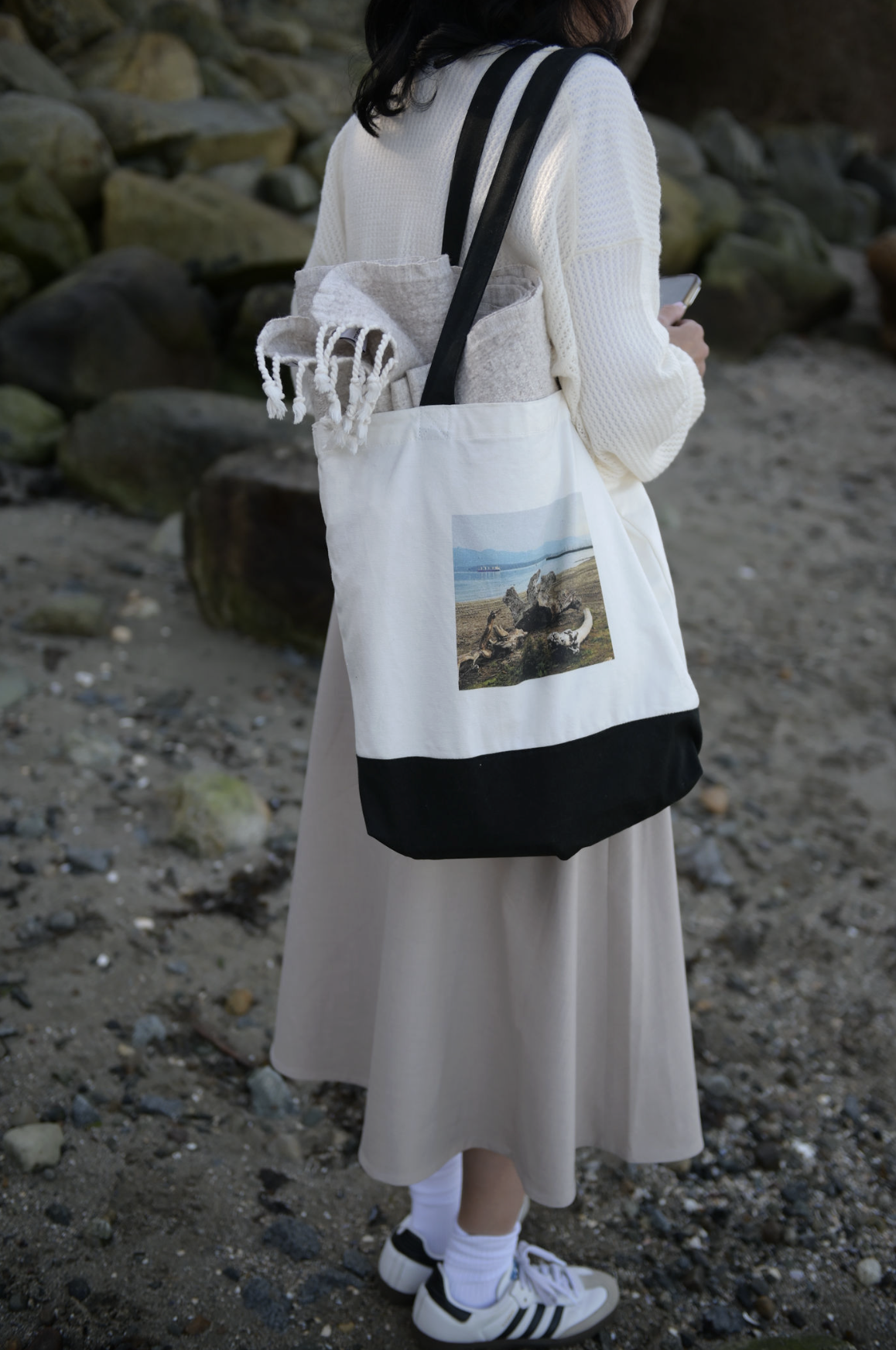 Person wearing a white sweater, beige skirt, and white sneakers with black stripes, carrying a tote bag with a beach scene. They are standing on rocky ground near large rocks, holding a phone.